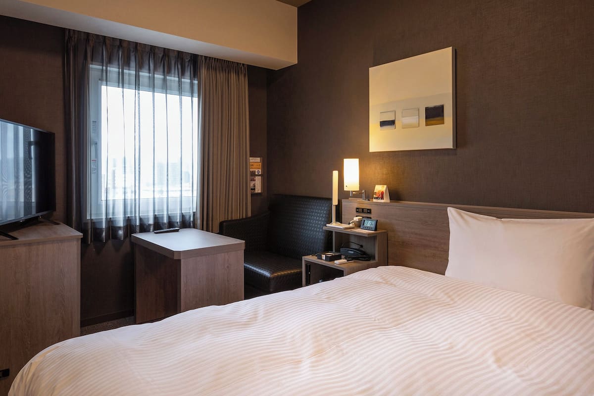 Habitación de hotel con cama, escritorio, silla, televisor y ventana con cortinas.