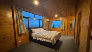 A wooden cabin bedroom with a bed, large windows, and a carpeted floor. - The Wood House Loft (Ngāruawāhia)