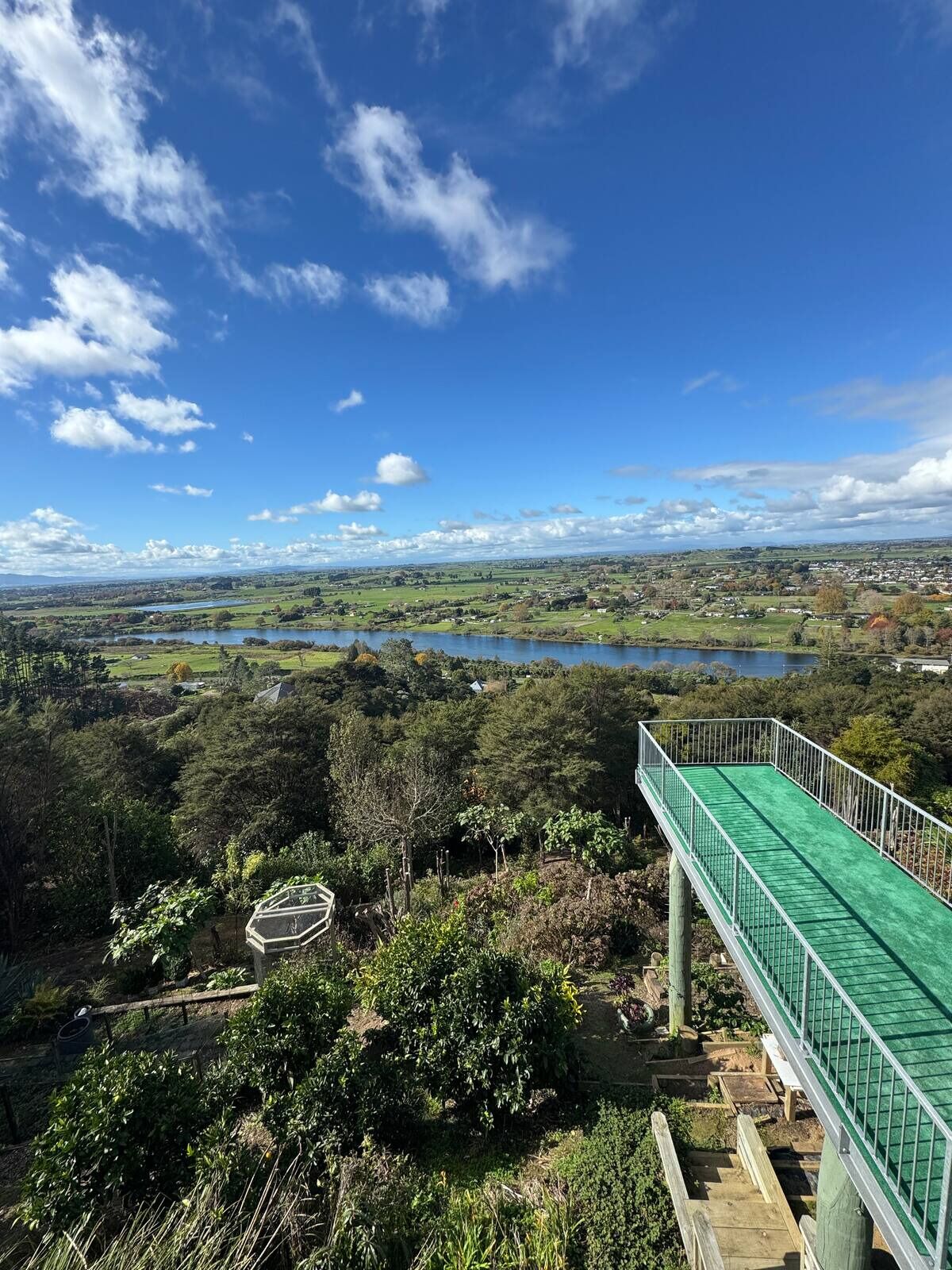 A scenic view from a high vantage point overlooking a lake, with a wooden deck and a green roofed structure in the foreground.