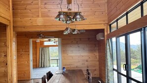 A wooden interior with a large dining table and chairs, a hanging light fixture, and large windows offering a view of the outdoors. - The Wood House Loft (Ngāruawāhia)