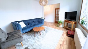 Una sala de estar con un sofá azul, una mesa de centro de madera, un televisor sobre un mueble de madera y un ventanal con plantas.