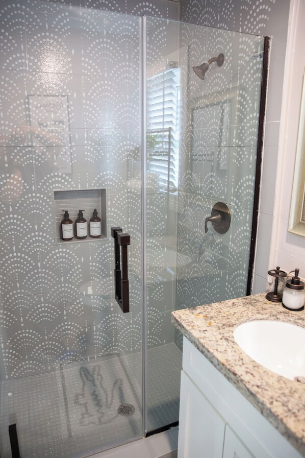 A modern bathroom with a glass shower enclosure, a built-in shelf with toiletries, and a granite countertop with a sink. - Westbury Getaway- PoolTable, Patio, &Grill- 4BD/3.5BA (Houston)
