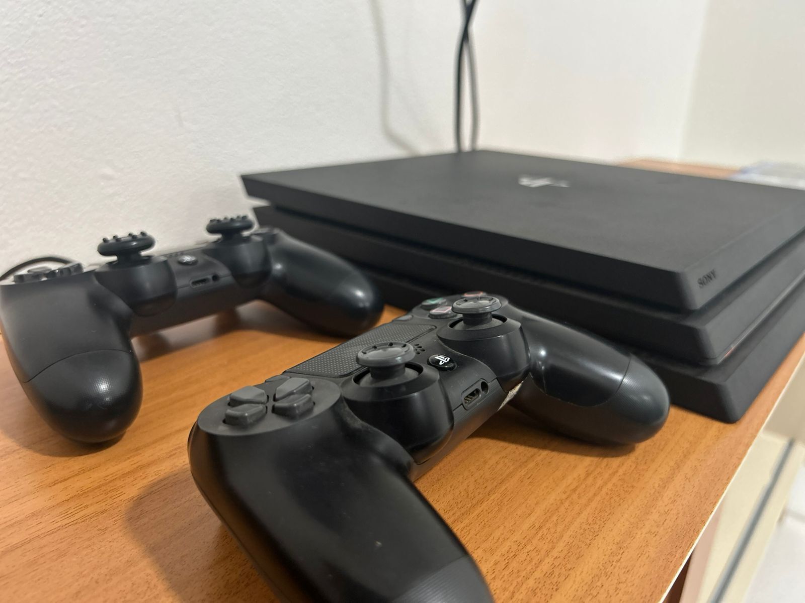 Two black game controllers on a wooden surface with a black electronic device in the background.