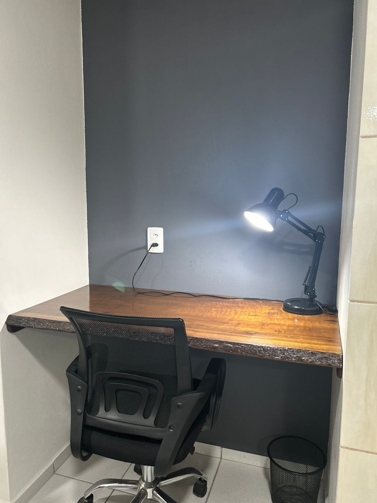 Un pequeño espacio de oficina con un escritorio de madera, una silla de oficina negra, una lámpara de escritorio y un cesto de basura.