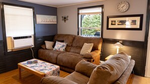 A cozy living room with a brown leather sofa, a wooden coffee table, and a side table with a lamp. There are two windows with blinds, a wall clock, and a framed picture. - Charming Cavendish-Area Cottage with River Views—Peaceful PEI Retreat (Prince Edward Island)
