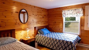 A wooden-paneled room with a bed, a nightstand with a lamp, a window with a floral valance, and a mirror on the wall. - Charming Cavendish-Area Cottage with River Views—Peaceful PEI Retreat (Prince Edward Island)