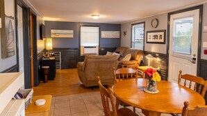 A cozy living room with a sofa, a dining table with chairs, a lamp, and framed pictures on the walls. - Charming Cavendish-Area Cottage with River Views—Peaceful PEI Retreat (Prince Edward Island)