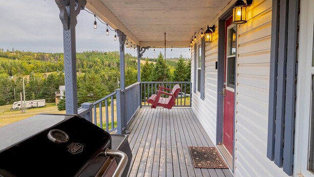 A covered porch with a grill, red chairs, and a view of a forested area. - Charming Cavendish-Area Cottage with River Views—Peaceful PEI Retreat (Prince Edward Island)