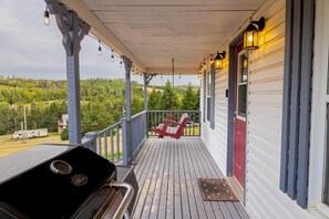A covered porch with a grill, red chairs, and a view of a forested area. - Charming Cavendish-Area Cottage with River Views—Peaceful PEI Retreat (Prince Edward Island)