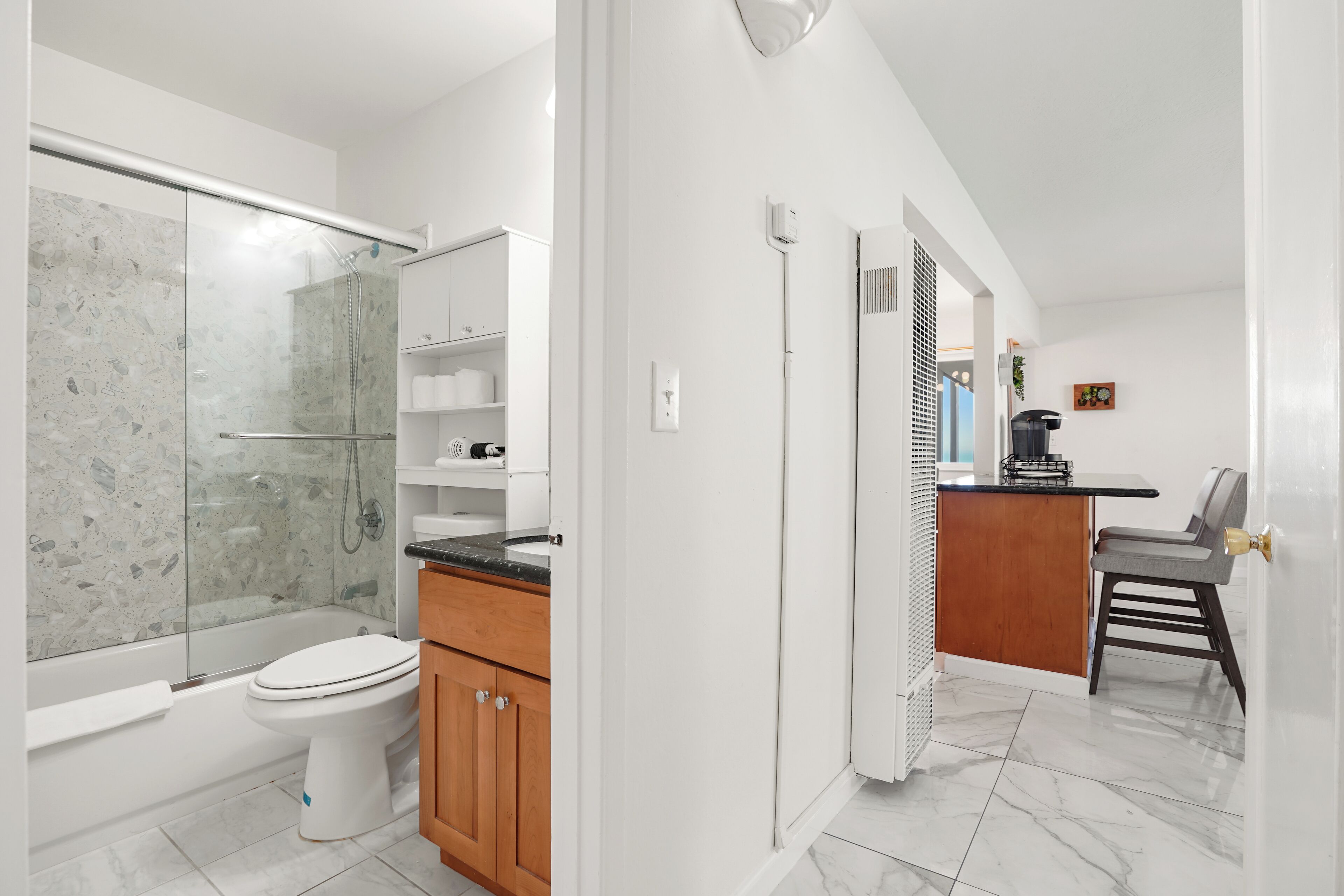 A modern bathroom with a glass-enclosed shower, a white toilet, wooden cabinets, and a small kitchenette area with a coffee maker.