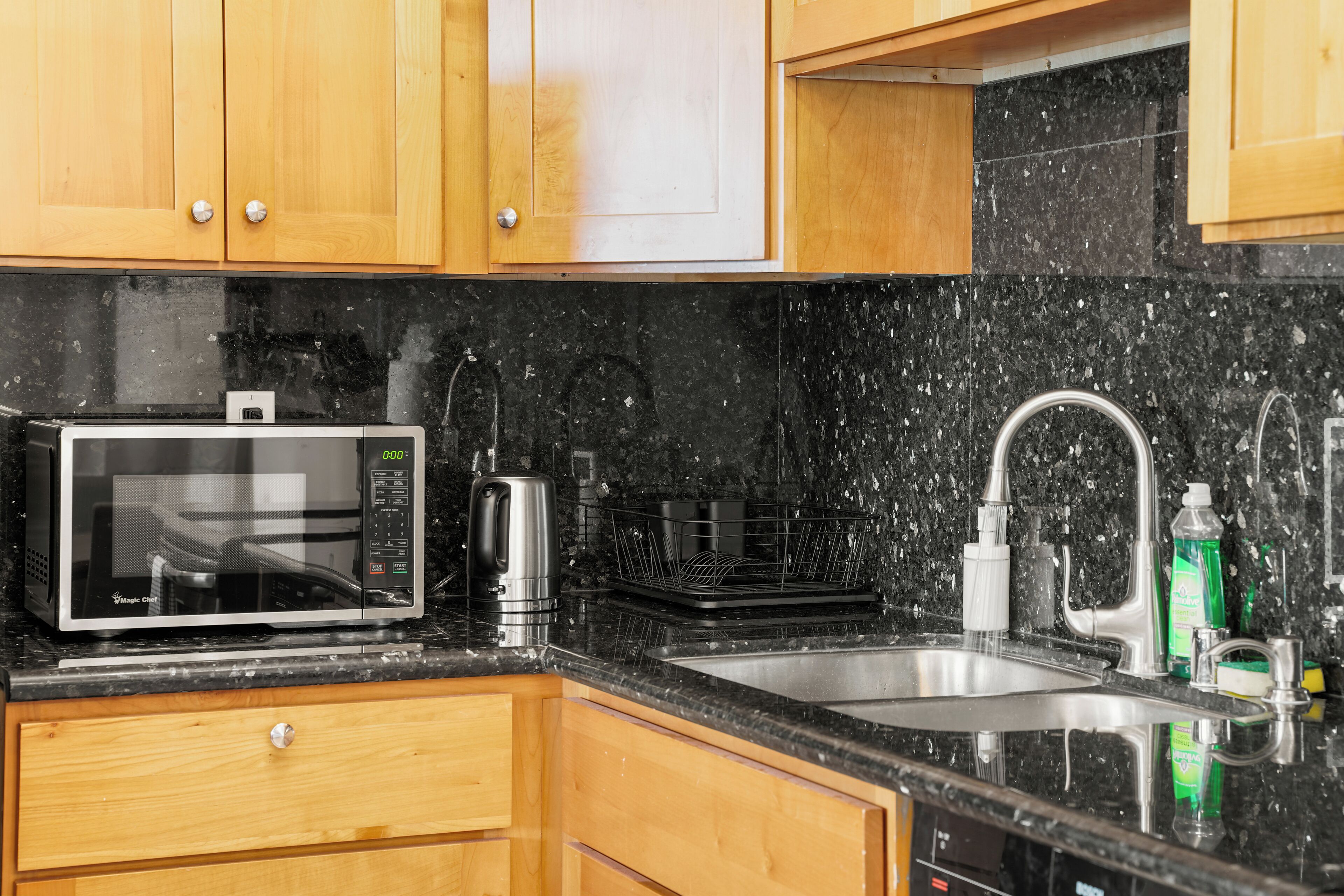 Een keuken met houten kastjes, een zwart granieten aanrecht, een roestvrijstalen spoelbak, een magnetron en een waterkoker.