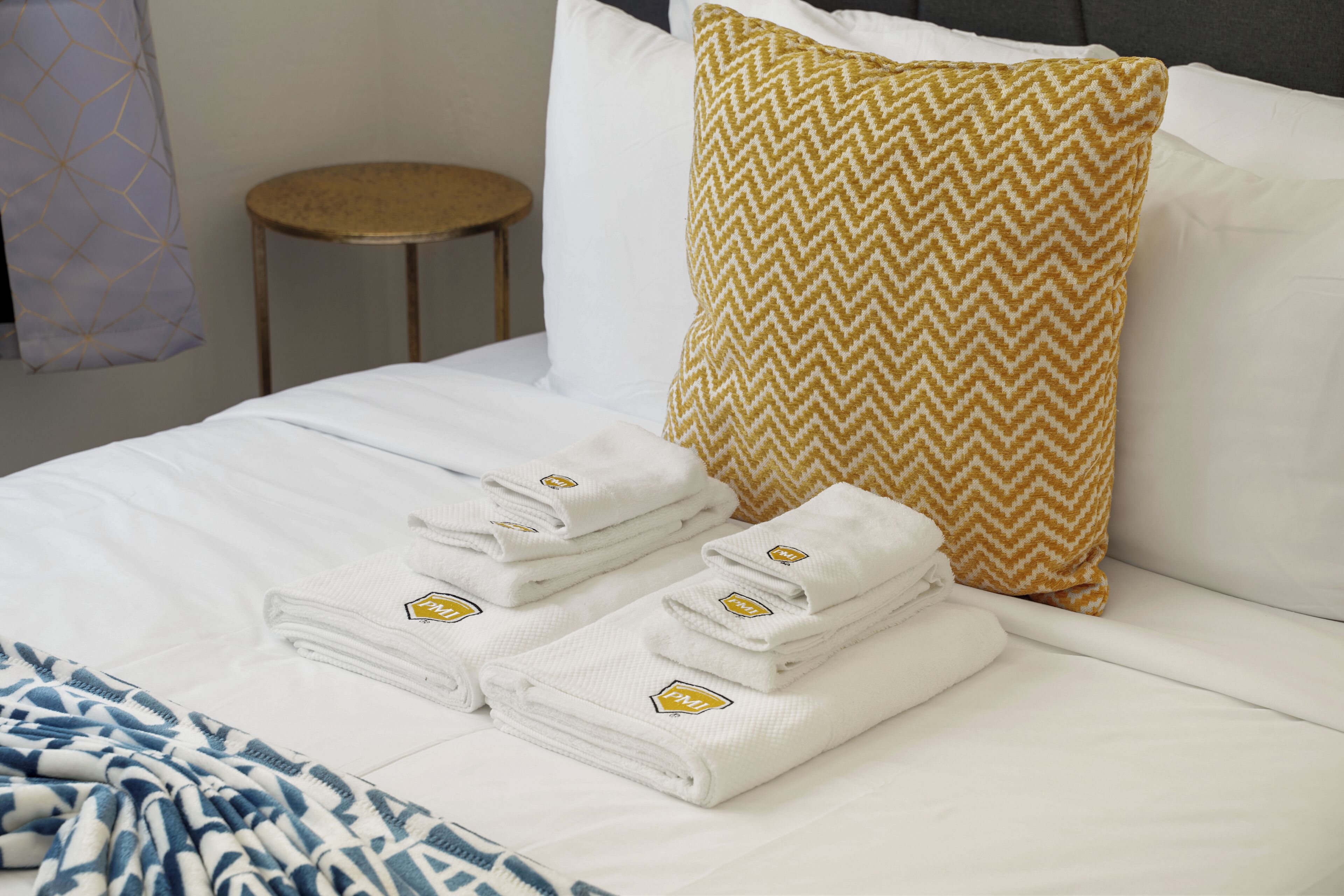 A neatly made bed with white linens and a folded towel set, a patterned pillow, and a small round stool.