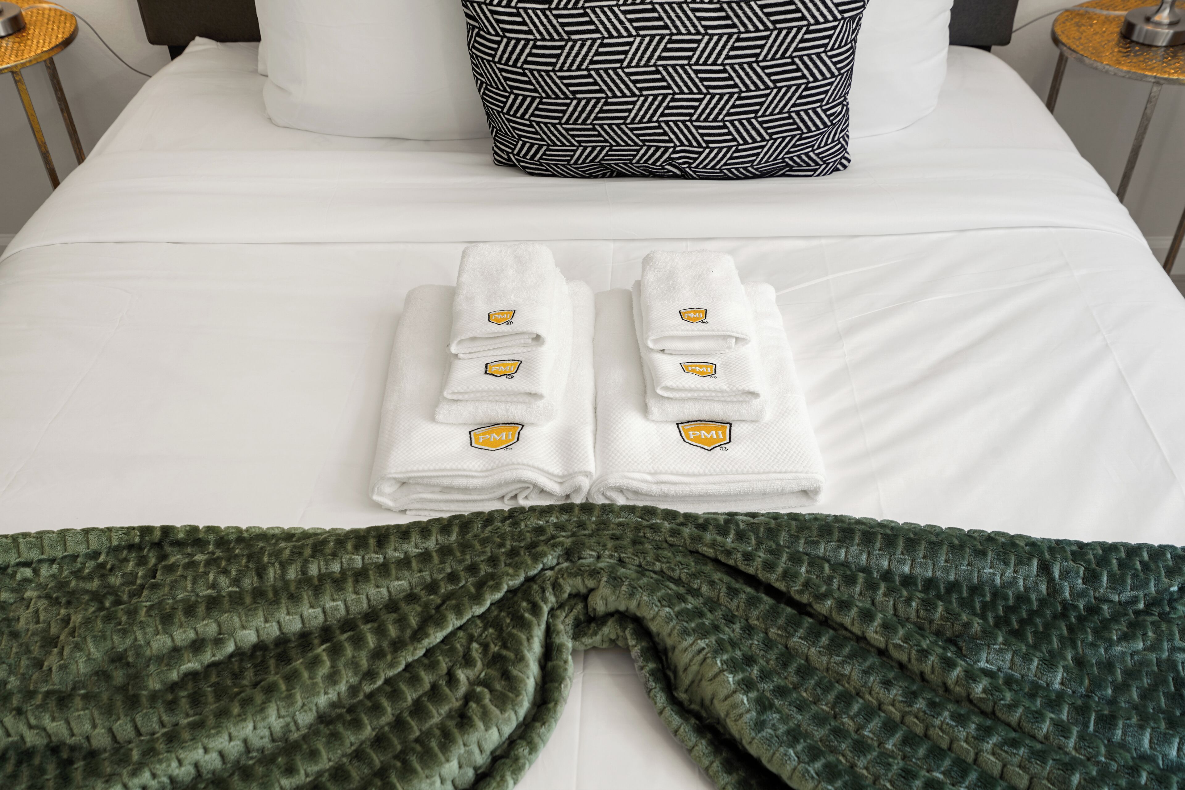 A neatly made bed with white sheets and two folded towels with a logo. A green textured blanket is draped over the bed.