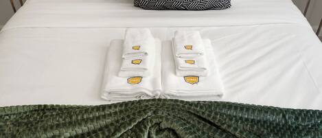 A neatly made bed with white sheets and two folded towels with a logo. A green textured blanket is draped over the bed.