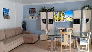 Una sala de estar moderna con una mesa de comedor de vidrio, un sofá color beige y un televisor de pantalla plana montado en la pared.