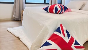 Ein Schlafzimmer mit einem Bett, zwei Kissen mit Union-Jack-Muster, beigen Vorhängen und Blick auf Gebäude durch große Fenster.