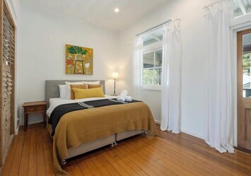 Unwind in this light-filled bedroom featuring a king bed, luxe linen, and warm timber floors. White curtains, soft lighting and calming tones create a peaceful space to rest after exploring Bangalow and Byron Bay.