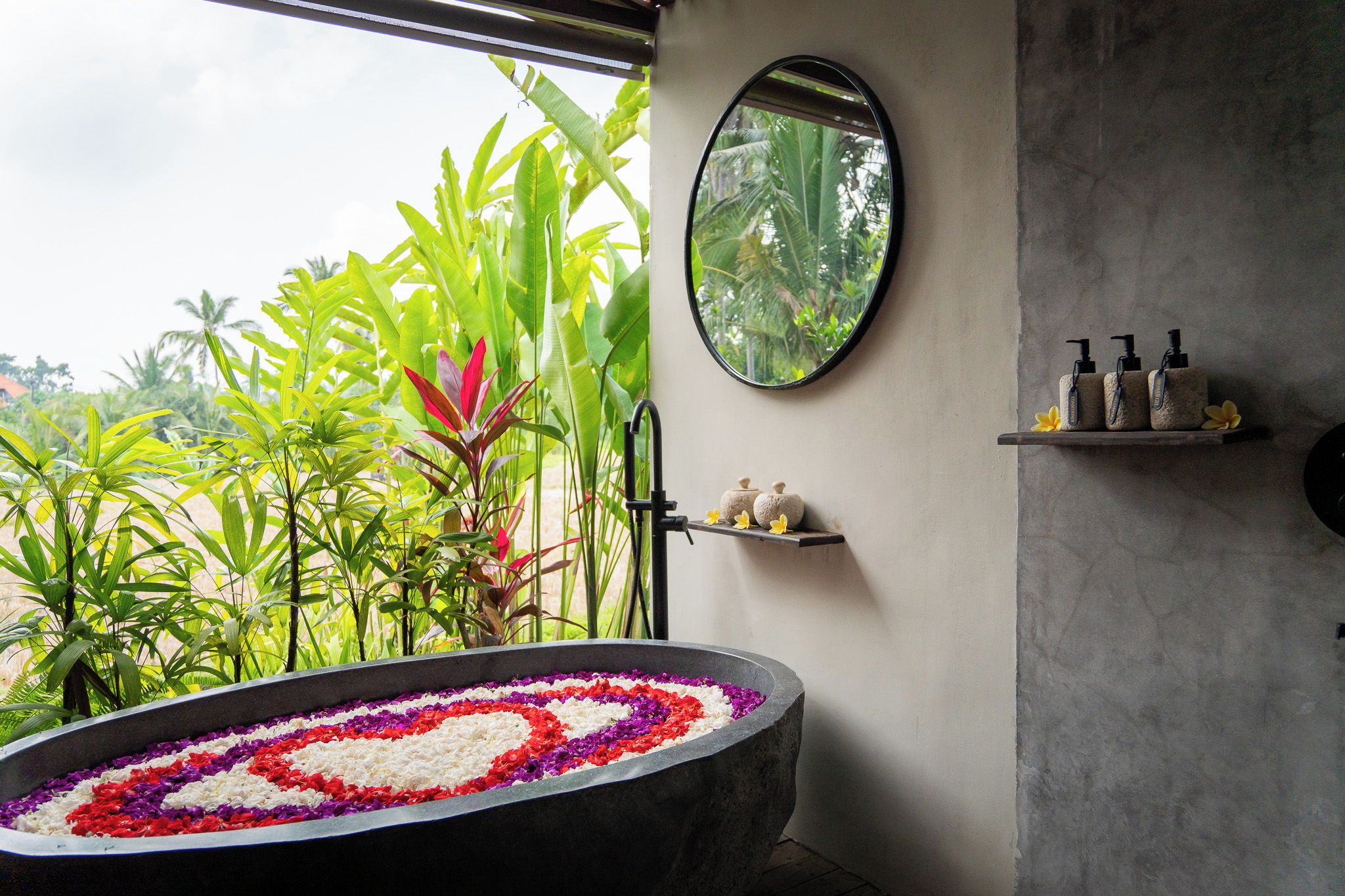 A bathroom with a large bathtub filled with flower petals, a round mirror, and a shelf with toiletries.