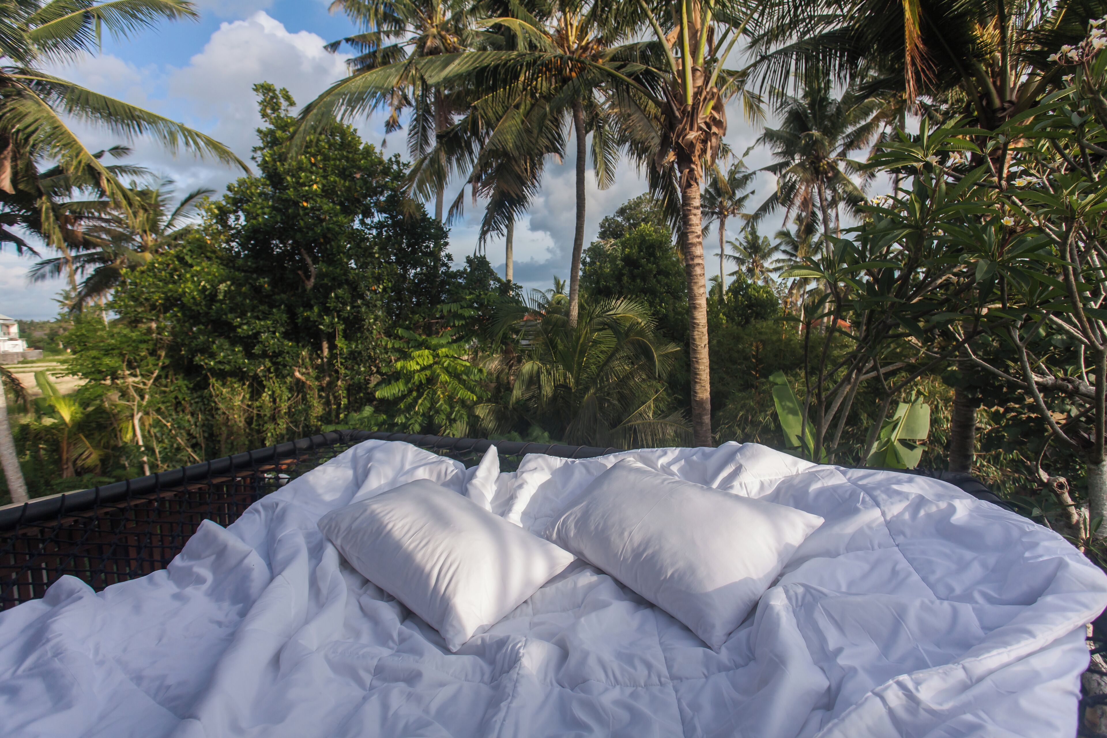 Una cama con ropa de cama blanca en una habitación con vista a la vegetación tropical.