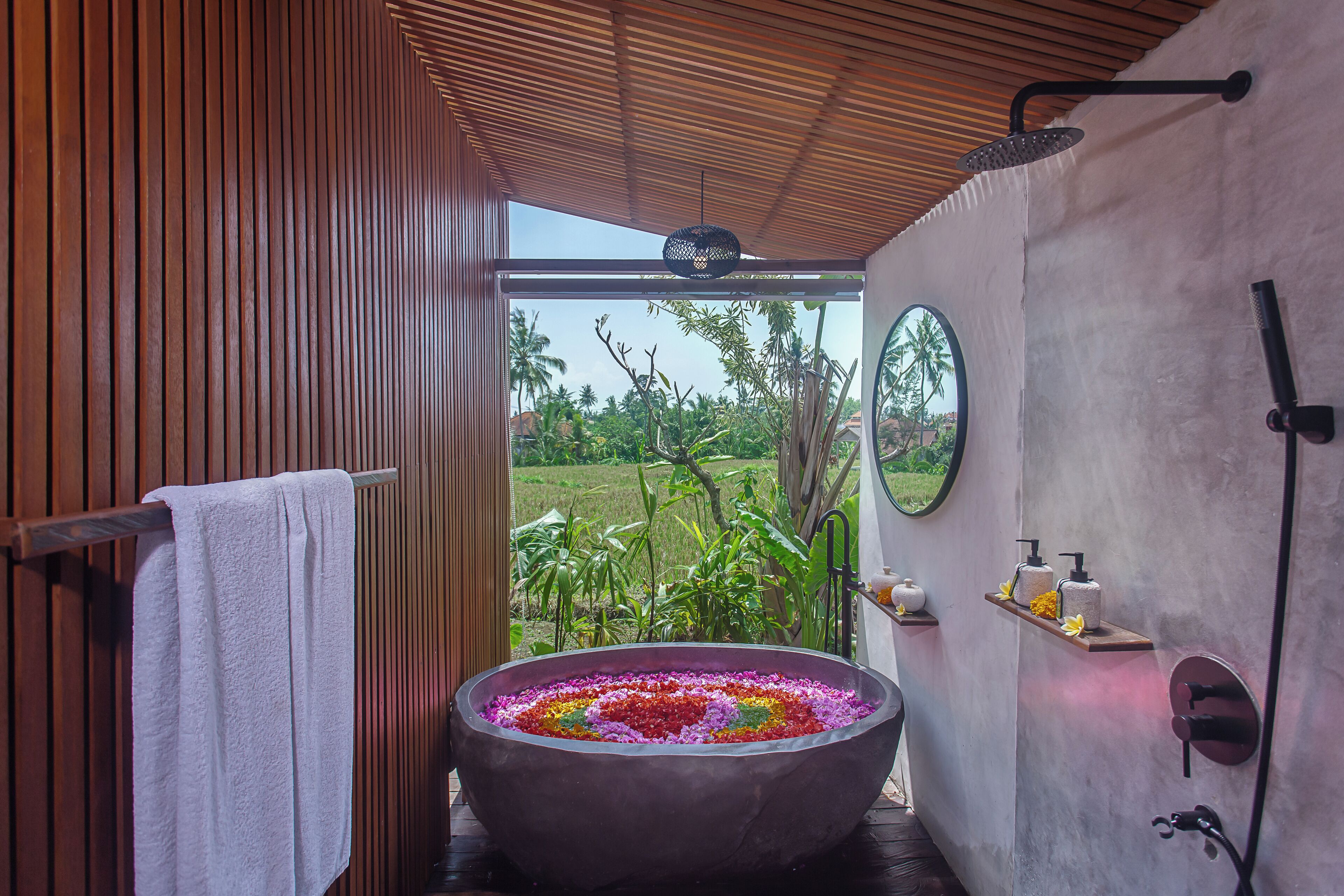 A bathroom with a large bathtub filled with flowers, a towel rack, a shower area, and a view of the outdoors.