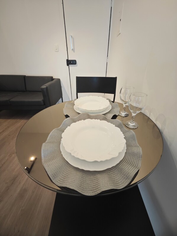 A dining table with two white plates and two wine glasses, set on a round table with a woven placemat. - Flat 209 norte Novo e Completo (Brasilia)