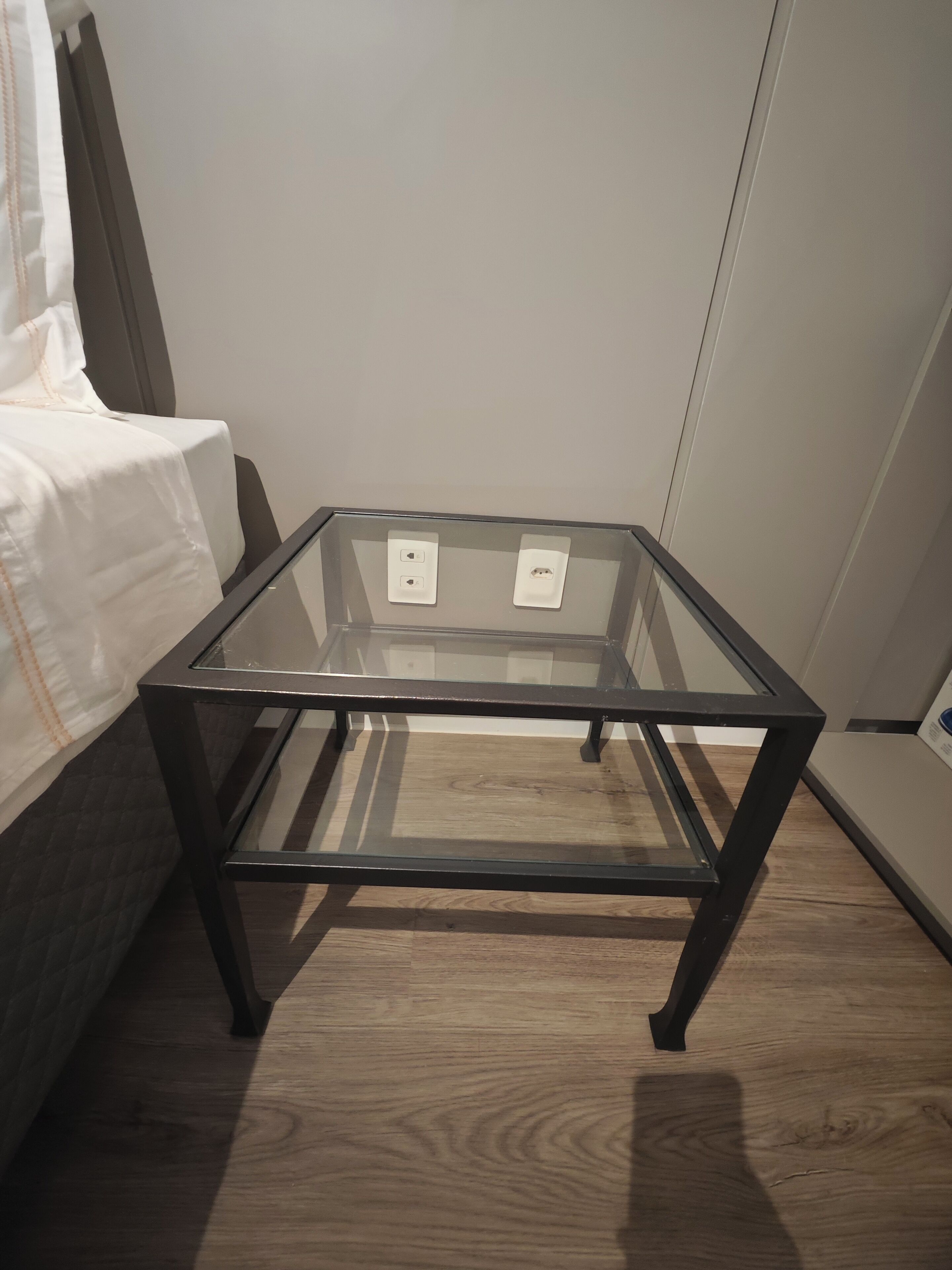 Una mesa con cubierta de vidrio y estructura de metal negro, dos enchufes y piso de madera.