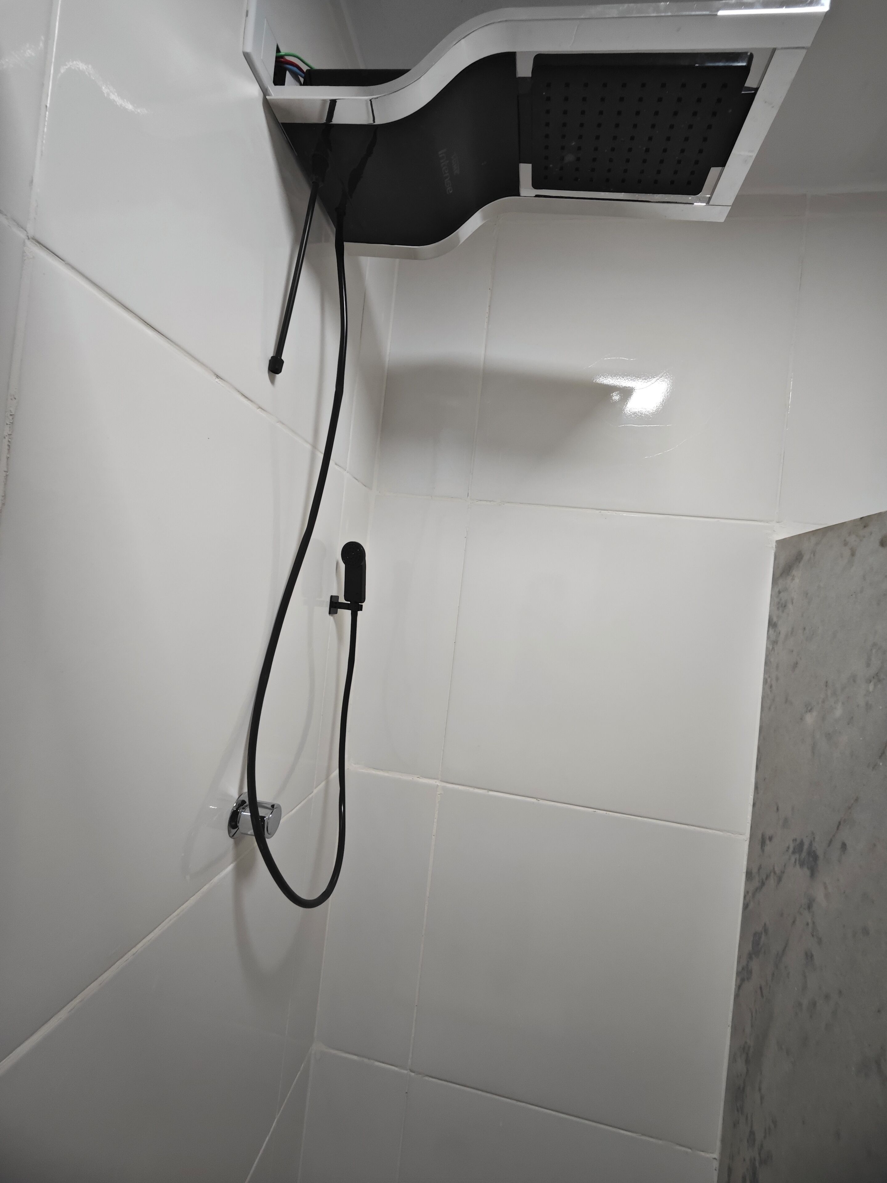 A corner of a bathroom with white tiles, a showerhead, and a black hose.