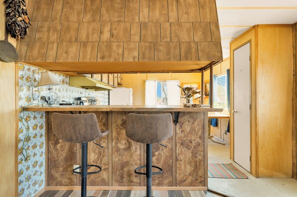 Una cocina con armarios de madera, una barra con dos taburetes, y una puerta visible que da a otra habitación.