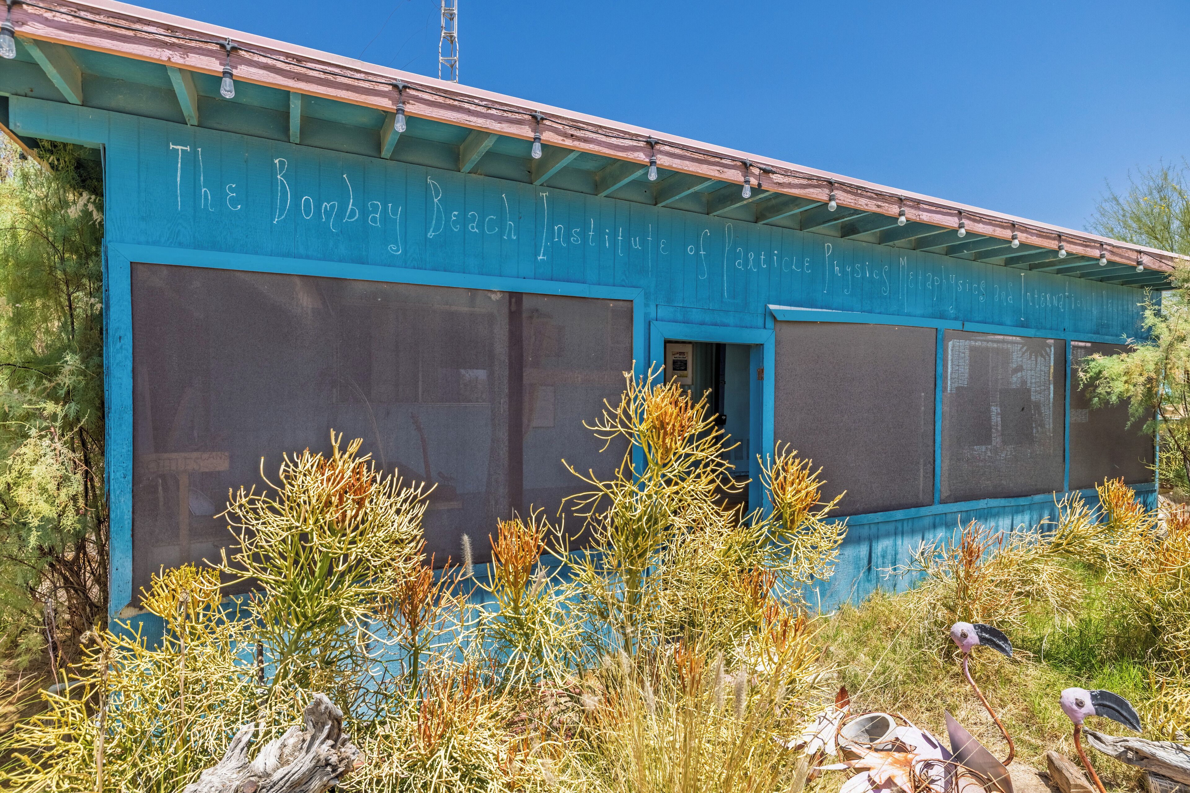 A building with large windows and a sign reading "The Bombay Beach Institute of Physical Metaphysics."