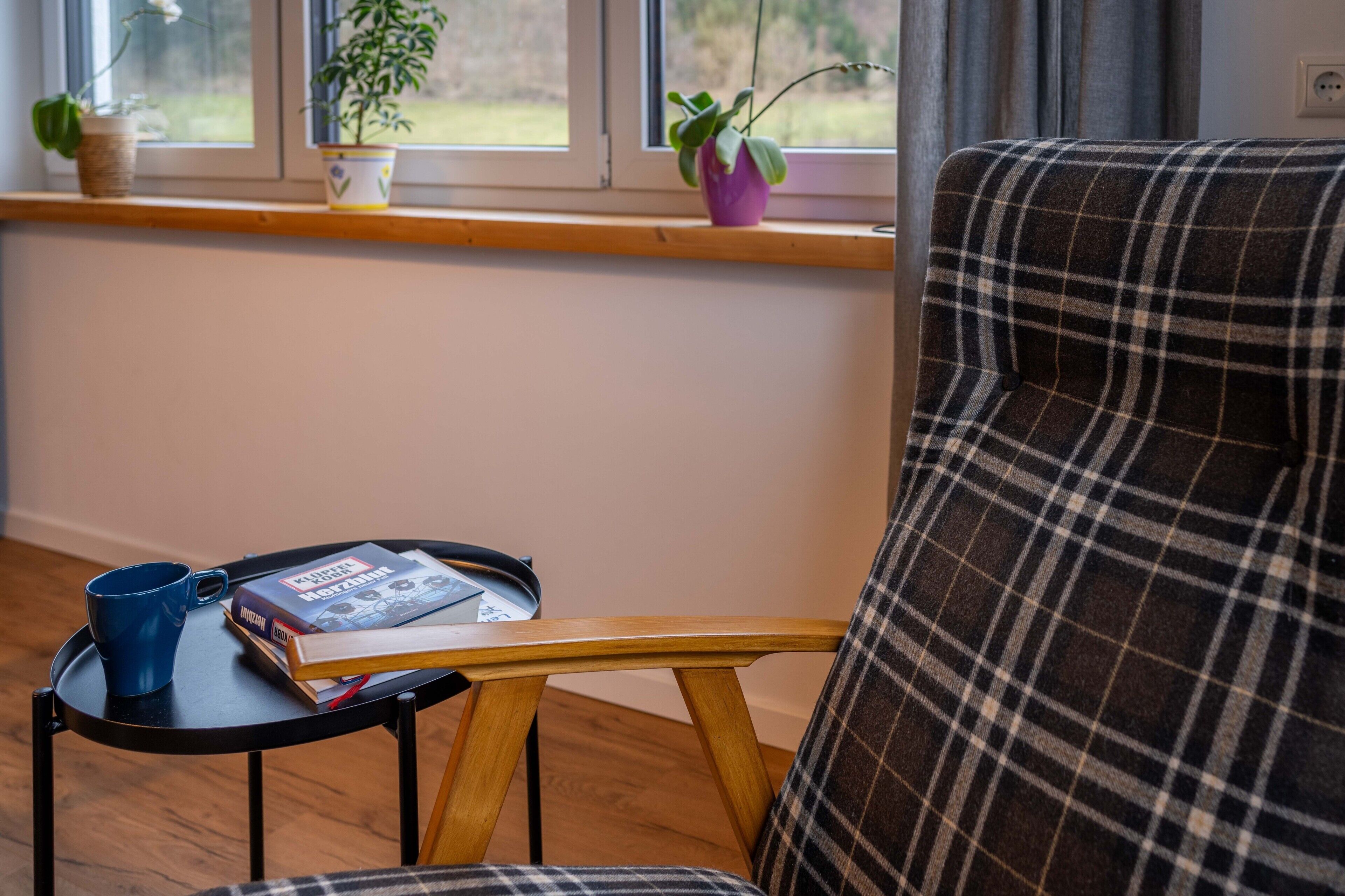Een kamer met een geruite stoel, een tafeltje met een blauwe mok en boeken, en een raam met planten.