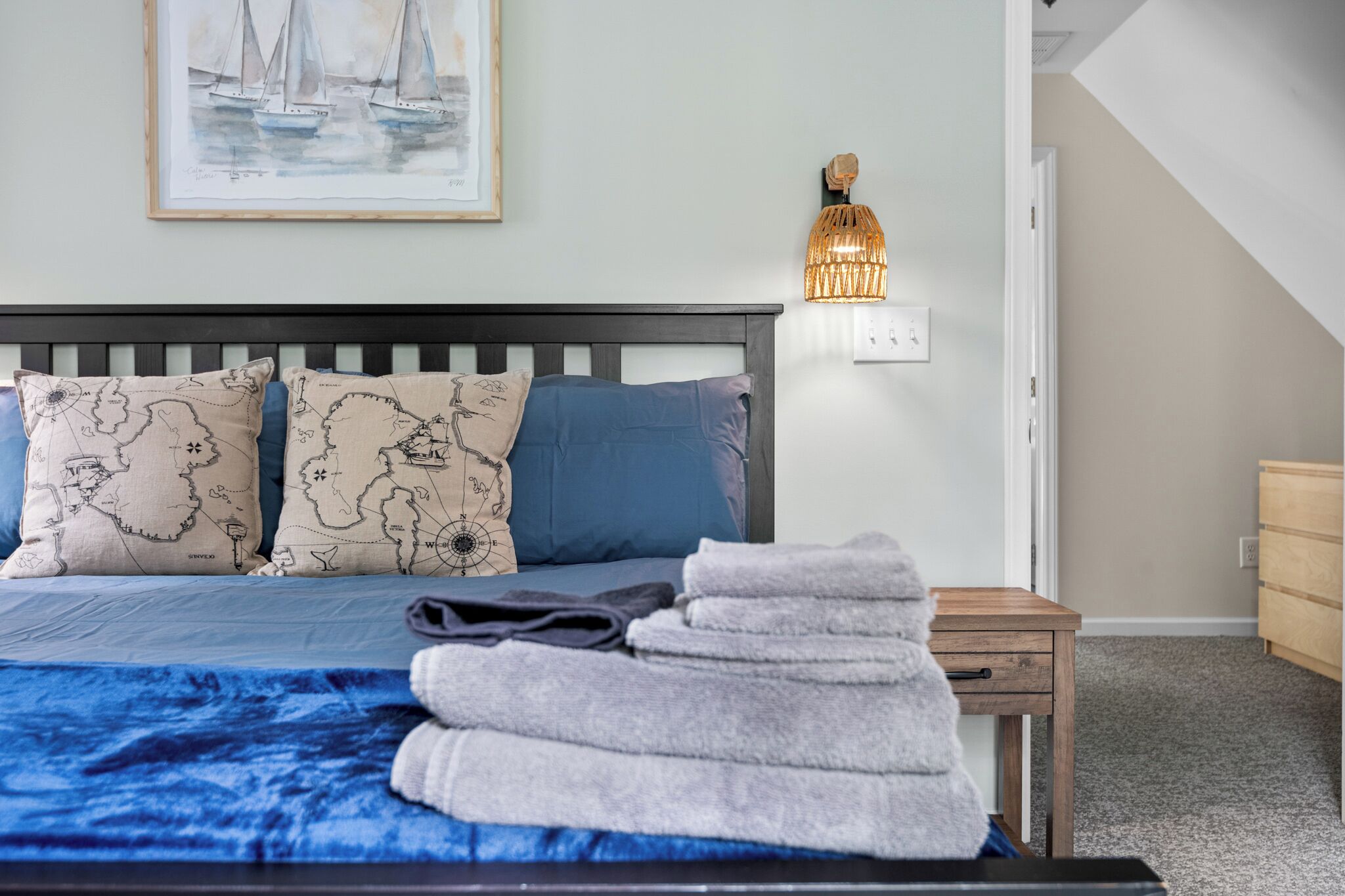 A neatly made bed with a blue blanket, two pillows with a map design, a small wooden nightstand with a lamp, and a framed painting of sailboats on the wall.
