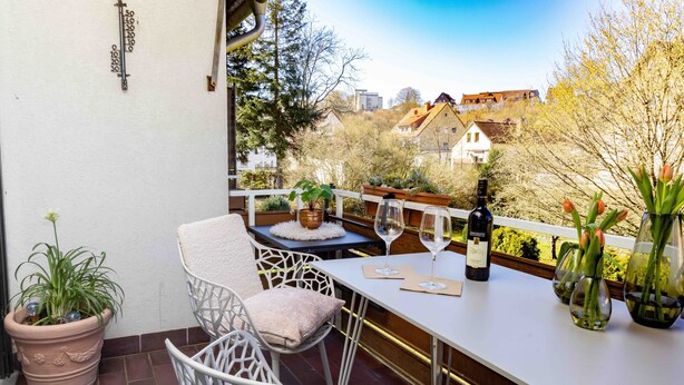 Ein Balkon mit Tisch, Stühlen, einem Blumentopf, einer Flasche und Weingläsern.