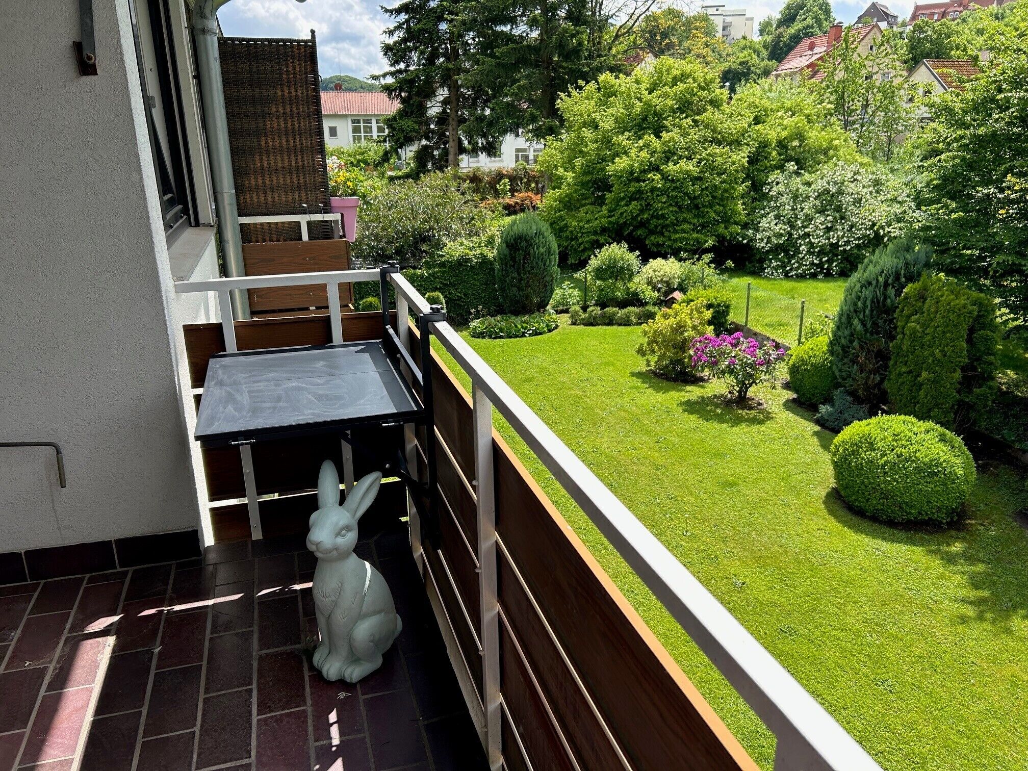 Un balcón con una pequeña mesa y una figurita de conejo blanco, con vista a un jardín cuidado con diversos arbustos y un césped impecable.