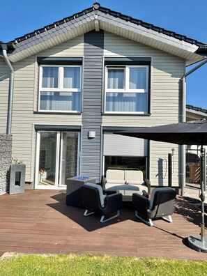 Ein zweistöckiges Haus mit einer Terrasse, die Gartenmöbel und eine Feuerstelle bietet.