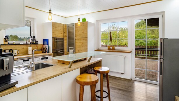 A modern kitchen with a wooden island, bar stools, a built-in oven, and a view of a garden through large windows.