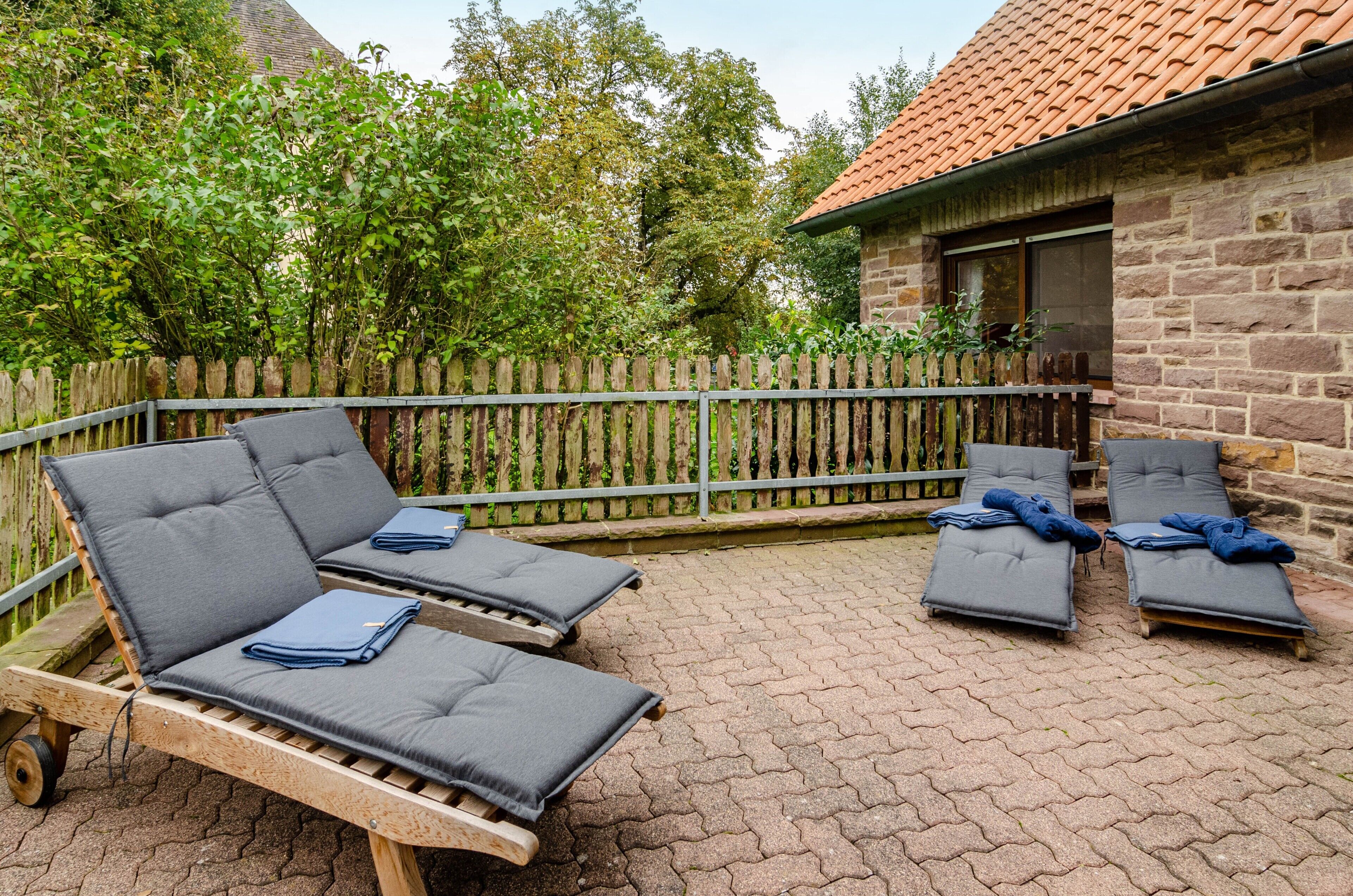 Eine Terrasse mit zwei Holzliegen und einem Steinhaus im Hintergrund.