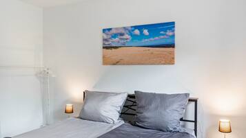 Ein Schlafzimmer mit einem Bett, zwei Nachttischlampen, einem Wandbild mit einer Strandansicht und einem weißen Heizkörper.