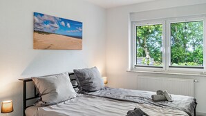 Une chambre à coucher avec un lit, des oreillers, une table de chevet avec une lampe, une fenêtre donnant sur des arbres et un tableau accroché au mur.