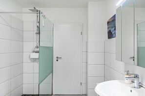 Un baño con cabina de ducha de vidrio, un lavamanos blanco y una puerta con panel de vidrio.