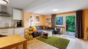 Una sala moderna con sofá, mesa de centro y mesa de comedor. Hay una cocina con armarios y estufa. Unos ventanales correderos dan a un balcón con vistas a la vegetación.