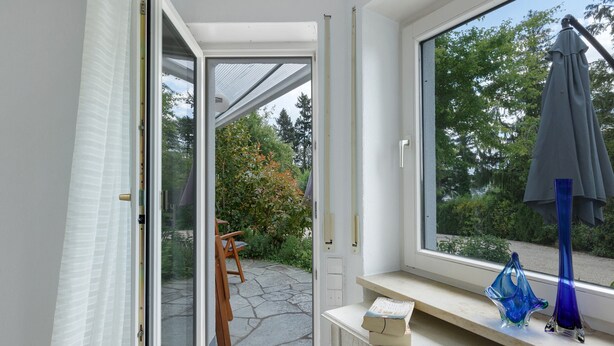 Una habitación con un ventanal grande, una silla de madera, una mesita y un jarrón azul.