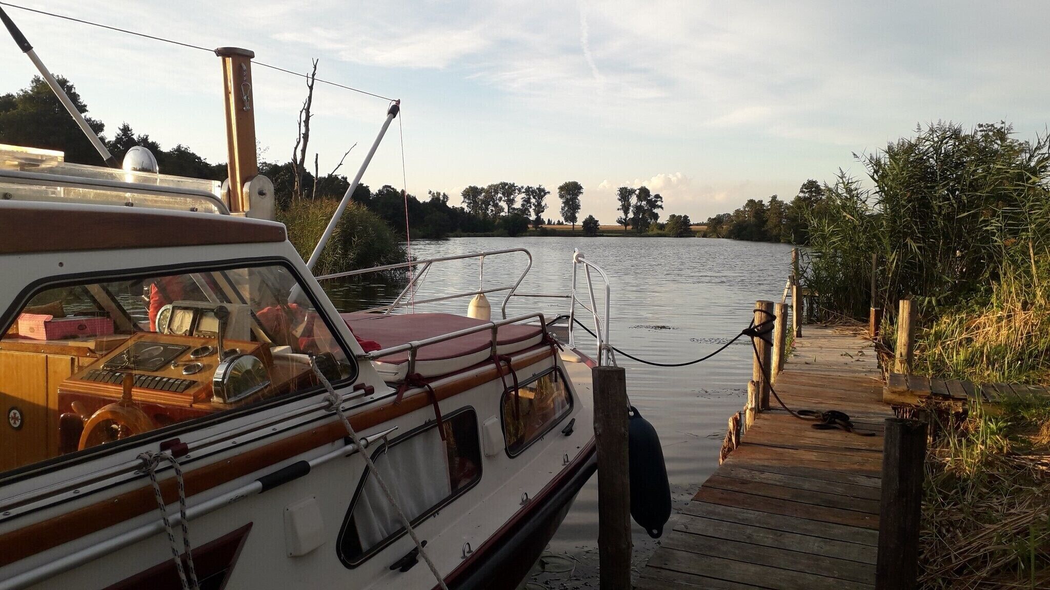 A boat docked at a wooden pier with a view of a calm river and trees in the background.
