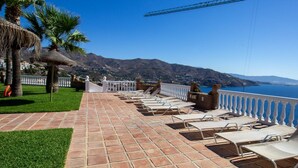 Een terras met lounge stoelen, een witte balustrade en uitzicht op zee en bergen.