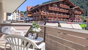 A balcony with white plastic chairs and a table, overlooking a wooden building with balconies and a mountain backdrop. - BeKb-Gebäude Studio No02 by Interhome (Wengen)