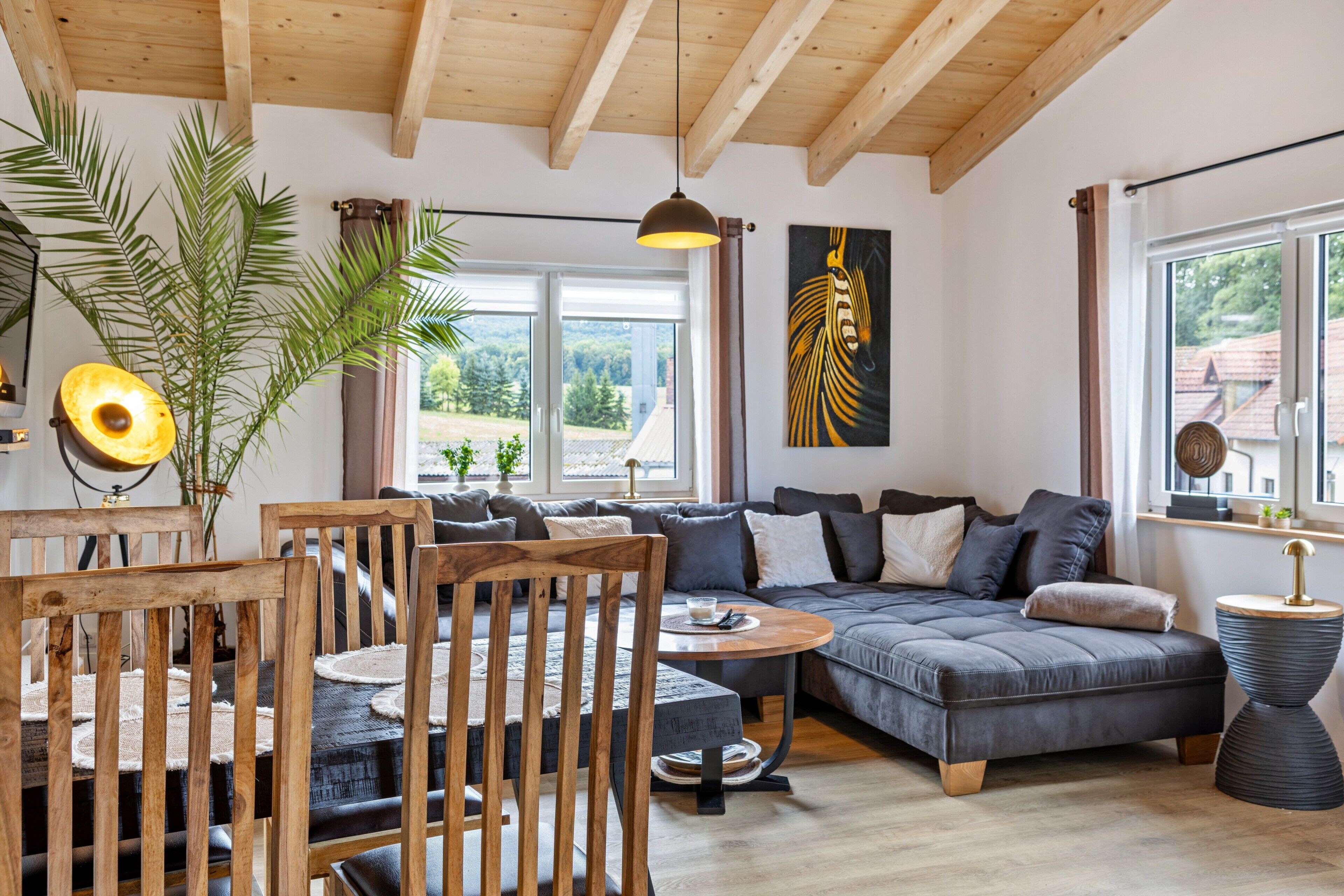 A modern living room with a wooden ceiling, a large window, a sofa, a dining table, and a plant.