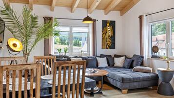 A modern living room with a wooden ceiling, a large window, a sofa, a dining table, and a plant.