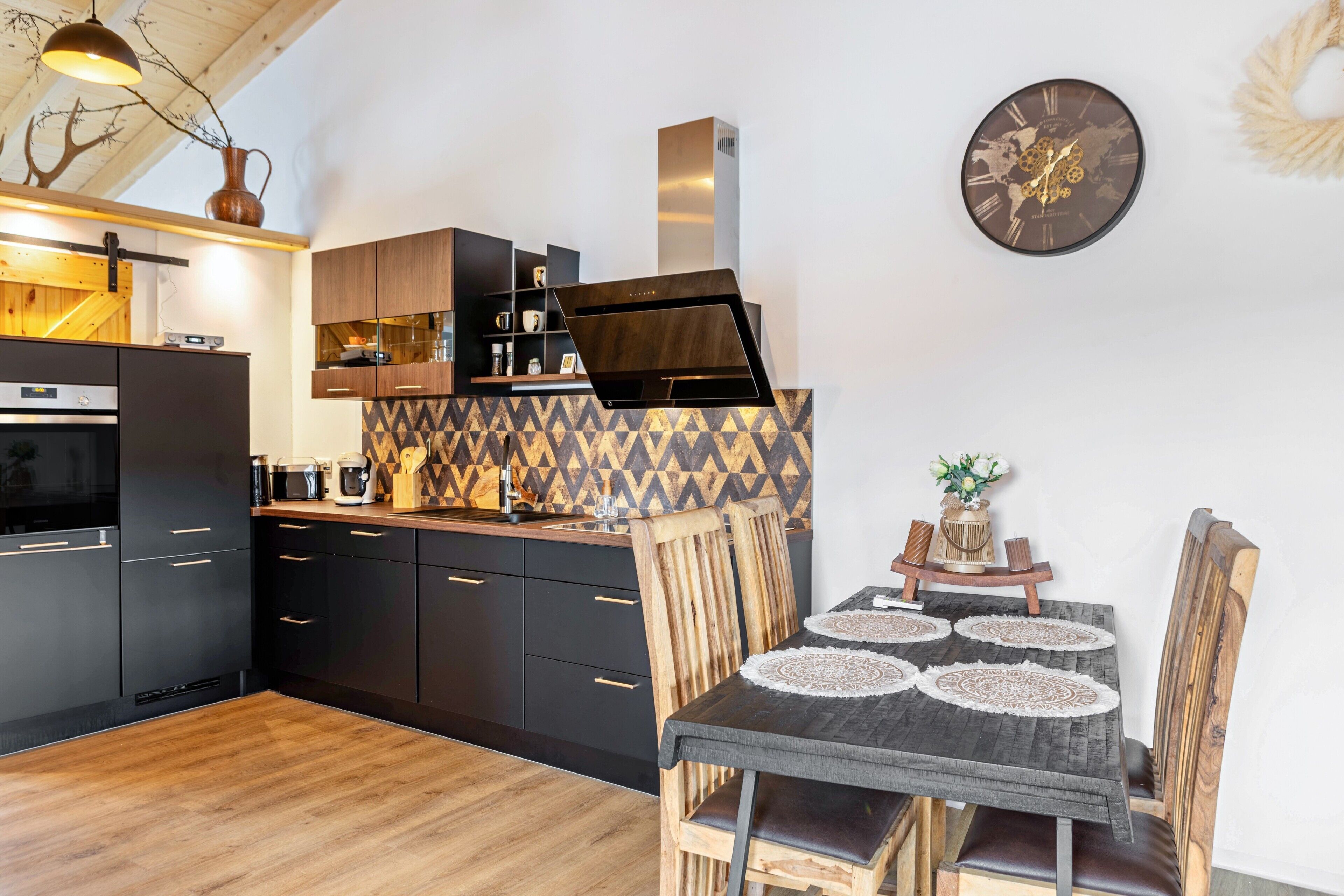A modern kitchen with dark cabinetry, a wooden dining table with a placemat, and a wall clock.
