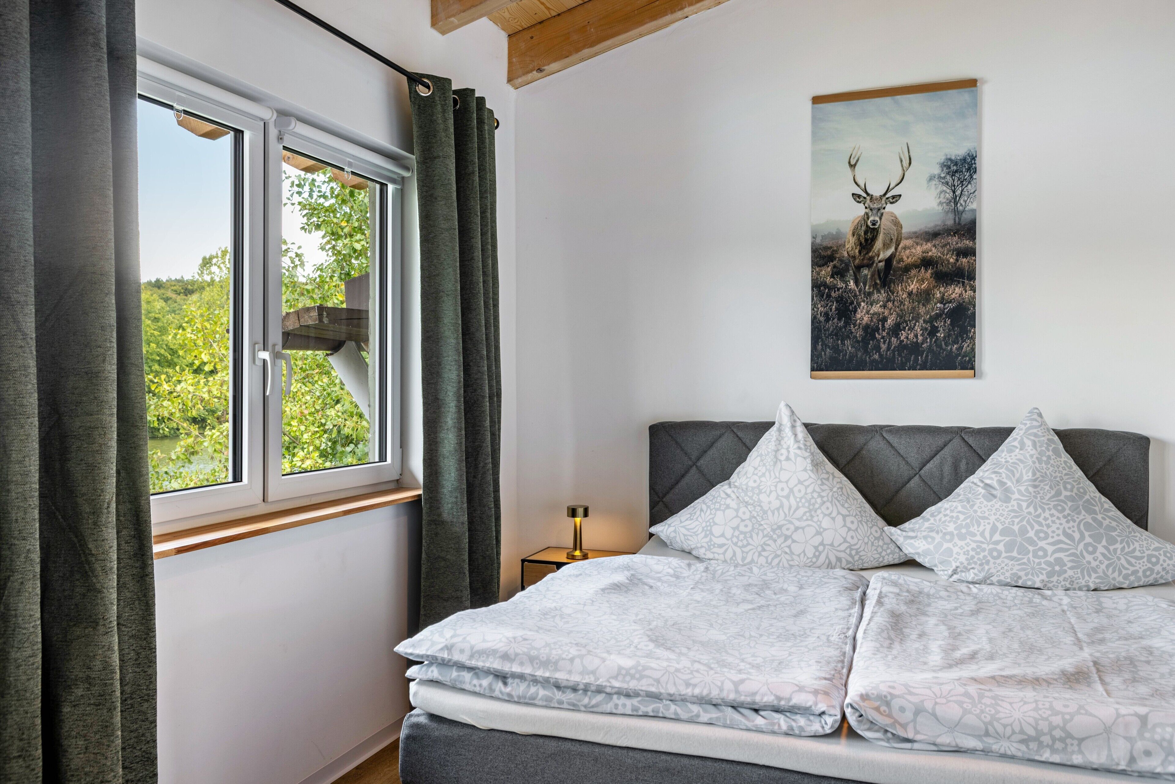 A bedroom with a bed, a window with curtains, a bedside table with a lamp, and a framed picture on the wall.