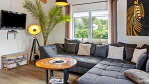 A modern living room with a sectional sofa, a round coffee table, a floor lamp, a television, and a large window with a view of greenery. - Holiday Apartment 'Im Garten Gottes' with Air Conditioning and Wi-Fi (Klosterlangheim)
