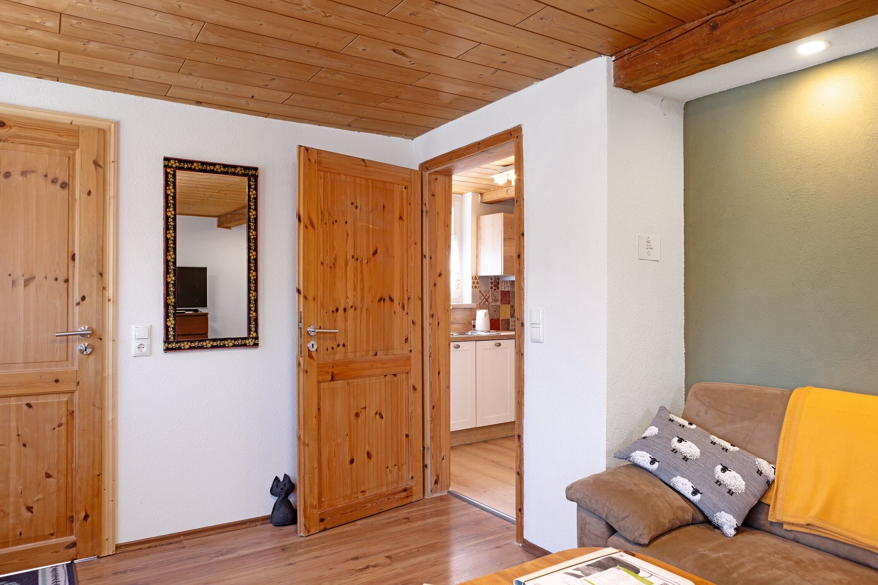 Una habitación con puertas de madera, un espejo, un sofá con un cojín estampado y una cocina al fondo.
